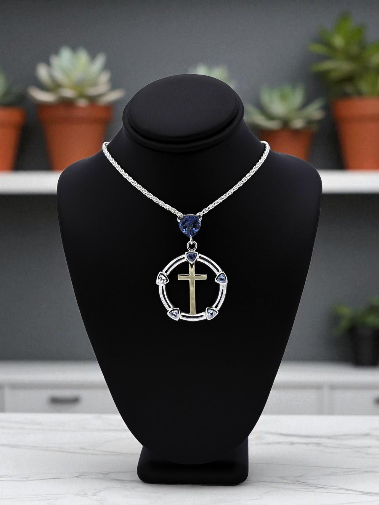 Necklace with a circular pendant featuring a cross and blue gemstone on a black bust, with a blurred indoor setting in the background.