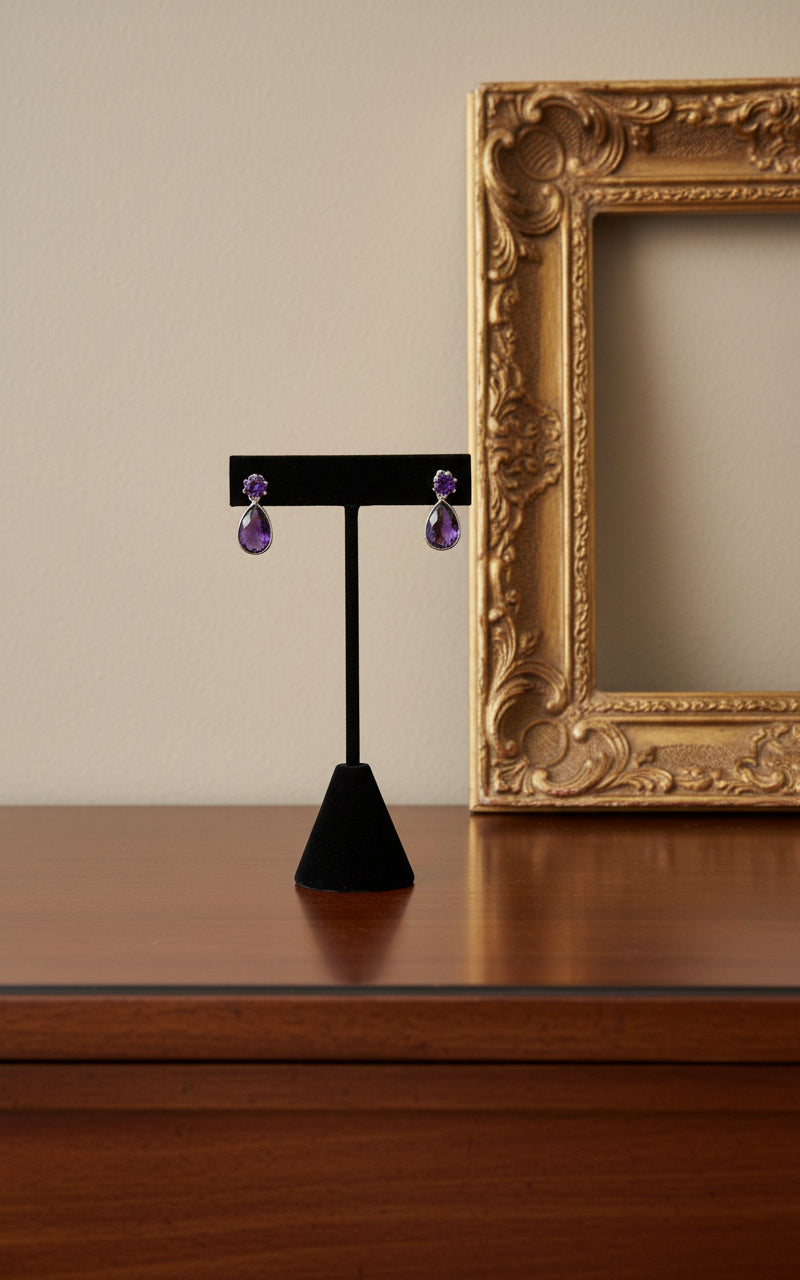 A pair of sterling silver dangle earrings featuring a large purple pear-shaped amethyst stone dangling from a smaller round amethyst. The earrings are displayed on a black stand.